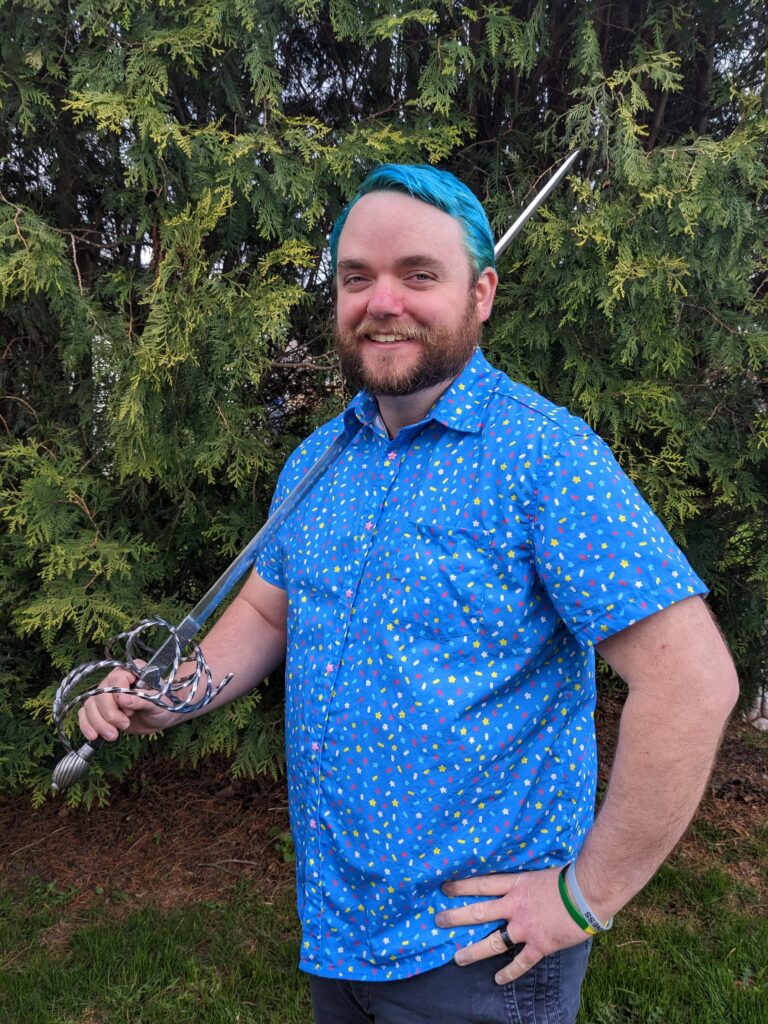 Author photo of Adam featuring a blue haired male wearing a bright blue shirt with a sprinkles pattern and holding a sword.